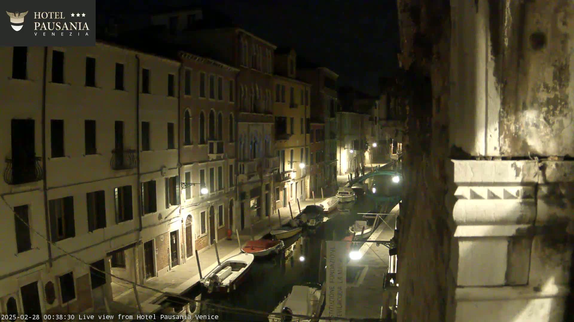 A nighttime view of a narrow canal in Venice, lined with buildings, with several boats moored along the waterway.