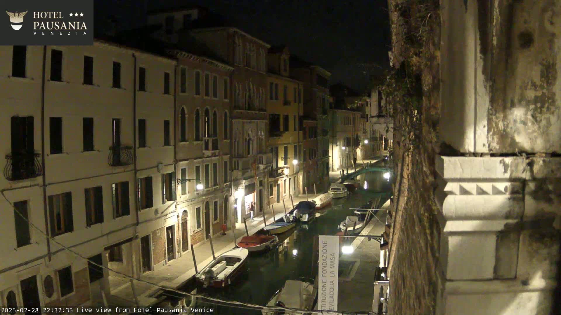 A nighttime view of a narrow canal in Venice, Italy, lined with buildings and several moored boats.
