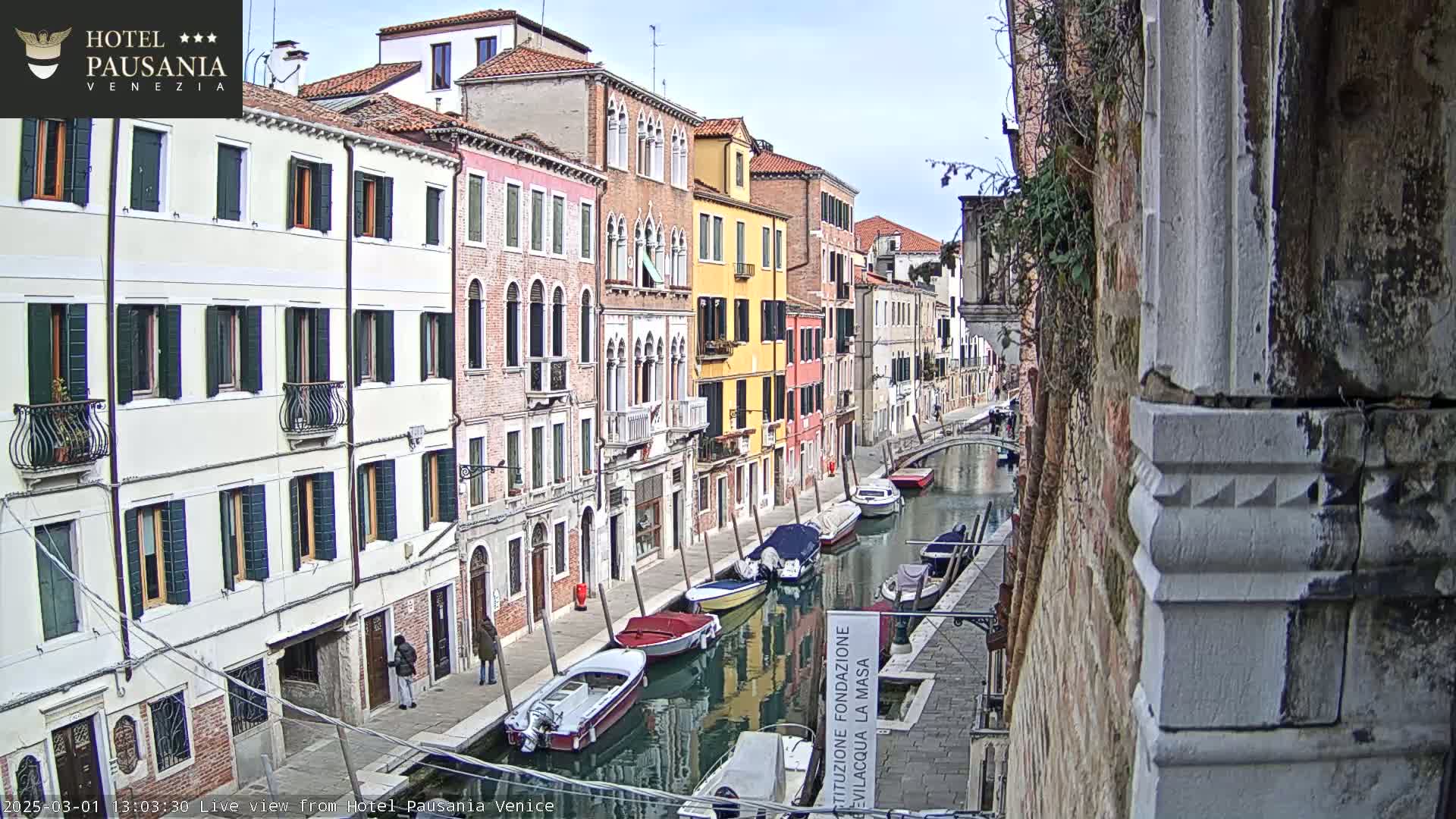 A canal in Venice, Italy, with colorful buildings lining its banks and several boats moored along the waterway, under a partly cloudy sky.