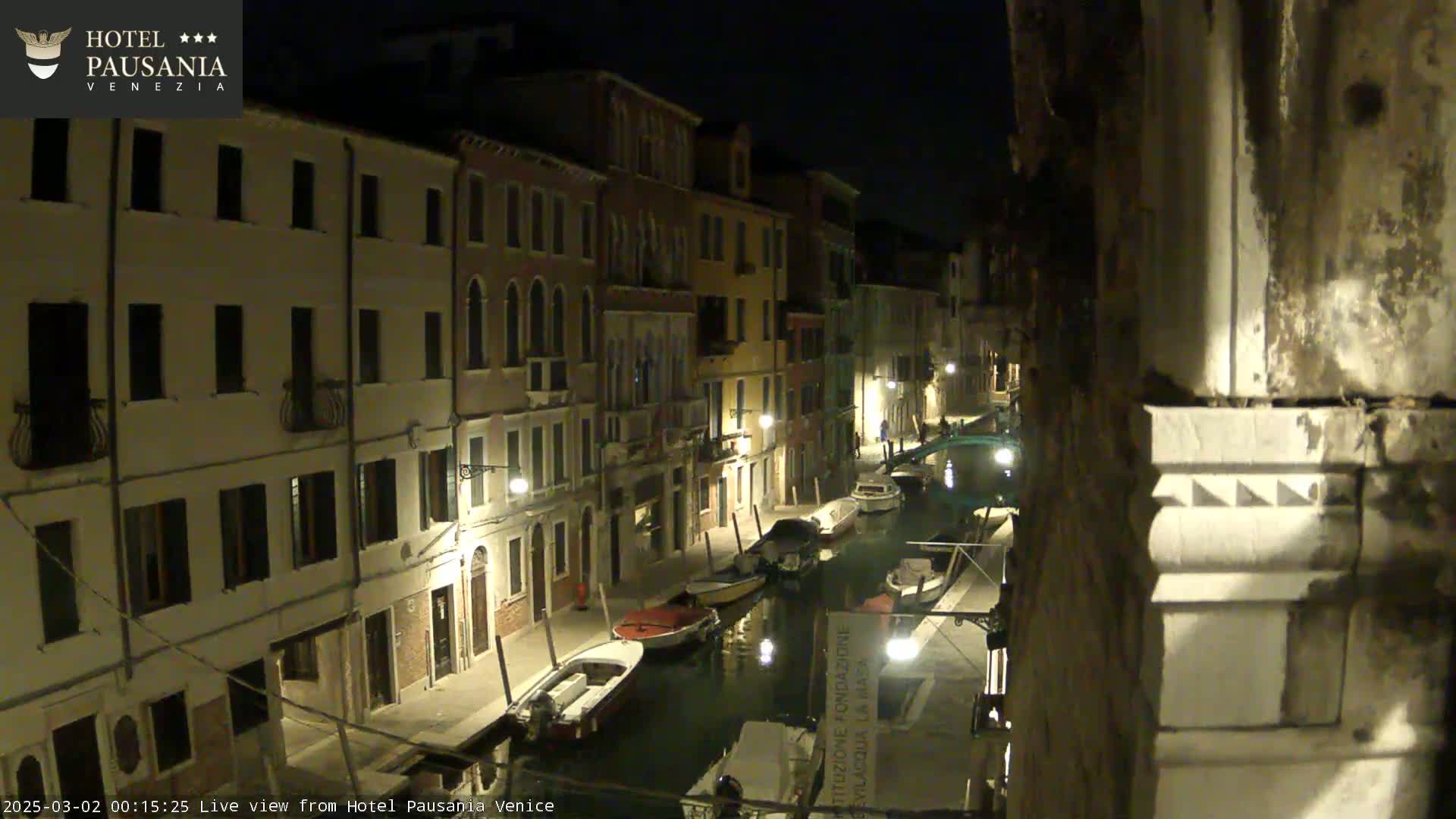 A nighttime view of a narrow canal in Venice, lined with buildings and several boats moored along its edges.