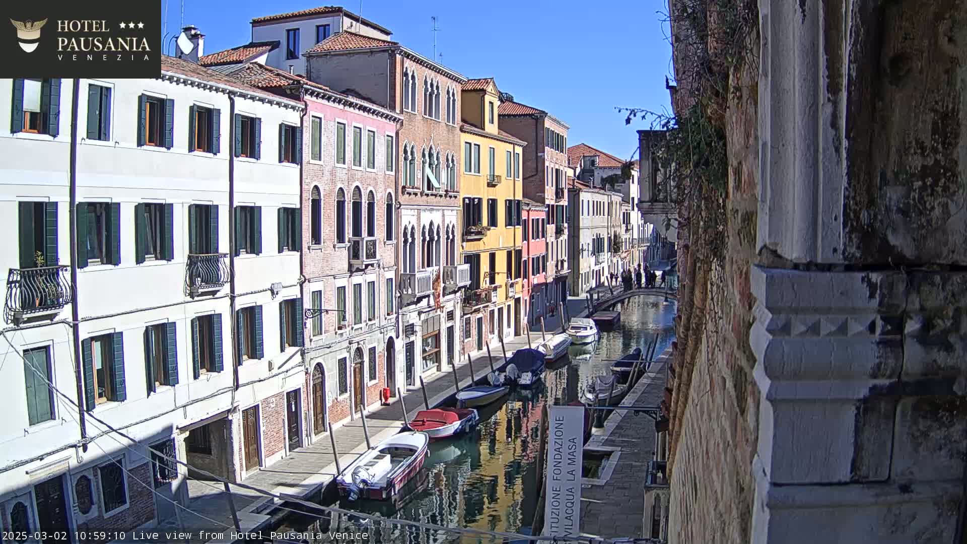 A sunny day in Venice shows a narrow canal lined with colorful buildings and several moored boats.