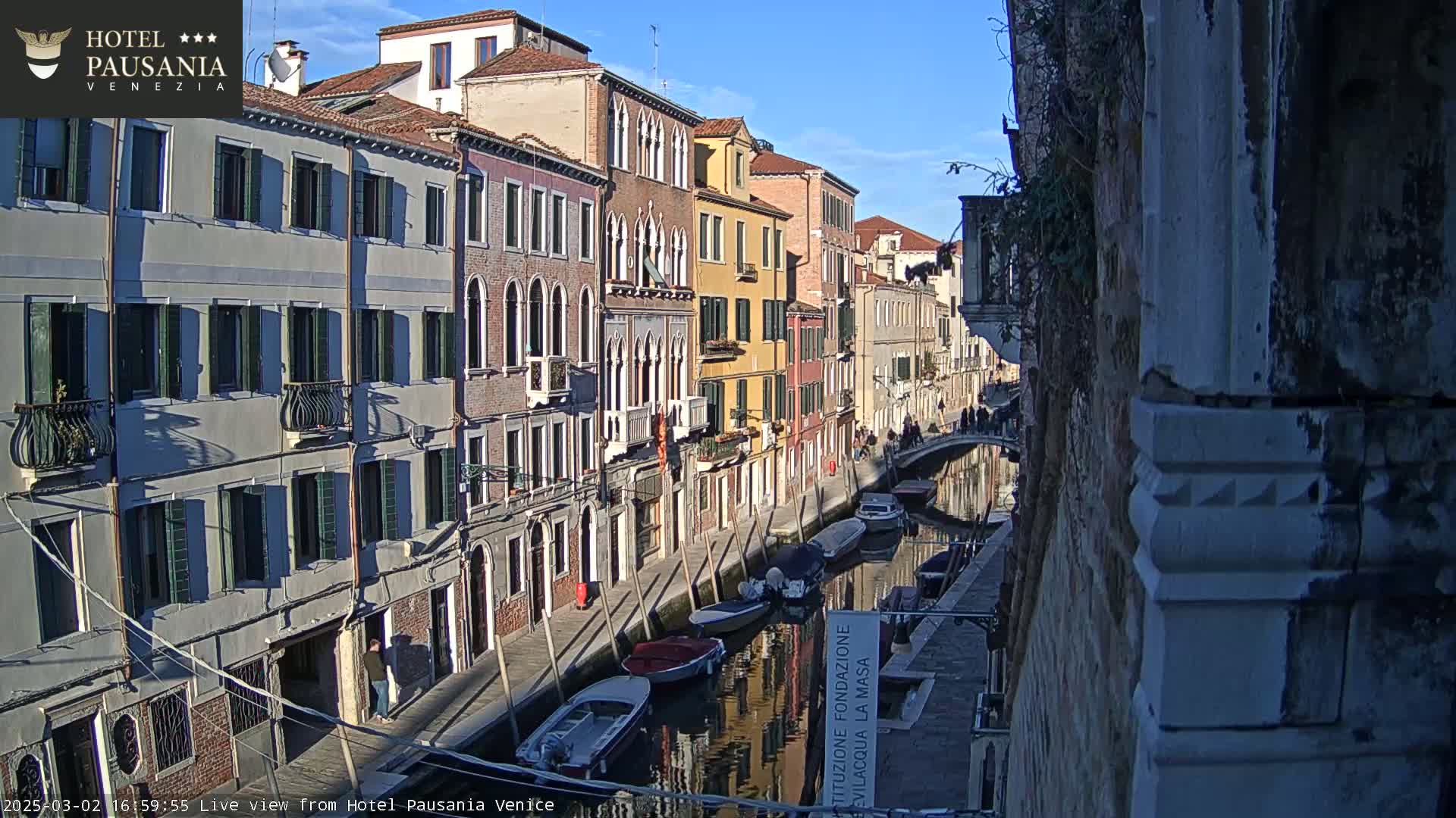 A sunny day in Venice shows a narrow canal lined with colorful buildings, several boats moored along the waterway, and pedestrians strolling on the canal-side walkway.