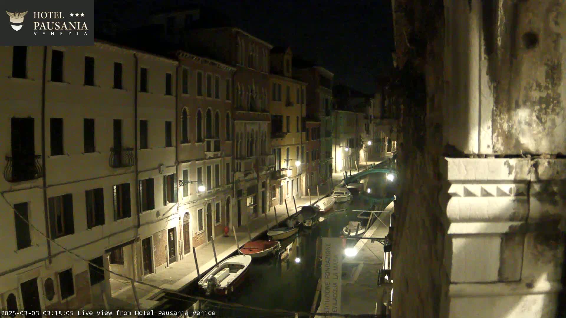A nighttime view of a canal in Venice, with several boats moored along its banks and illuminated buildings lining the waterway.