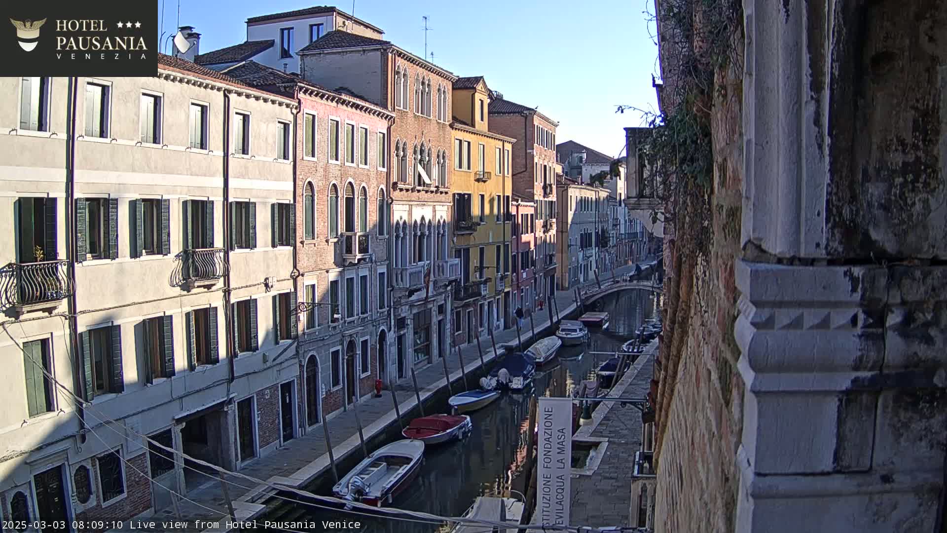 A sunny view of a Venetian canal lined with colorful buildings and several moored boats.