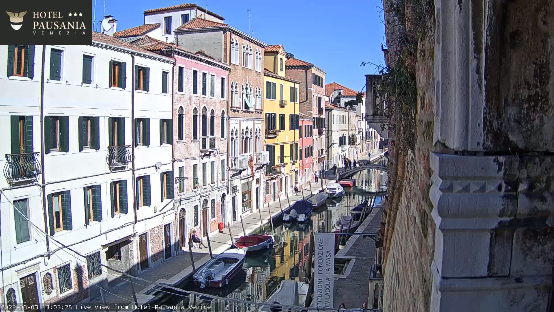 A sunny day in Venice shows colorful buildings lining a canal with several boats moored along the waterway.