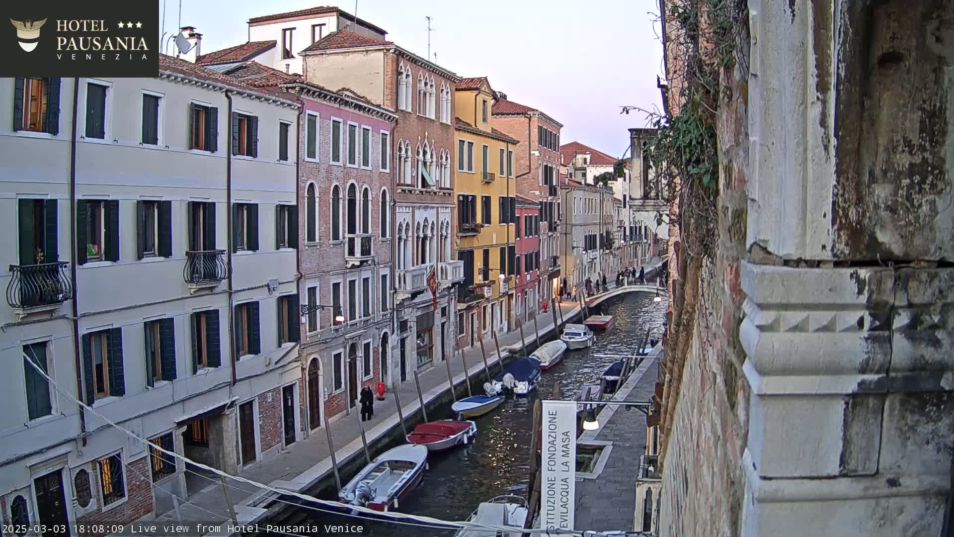A narrow canal in Venice, lined with tall, colorful buildings, is filled with several moored boats under a twilight sky.