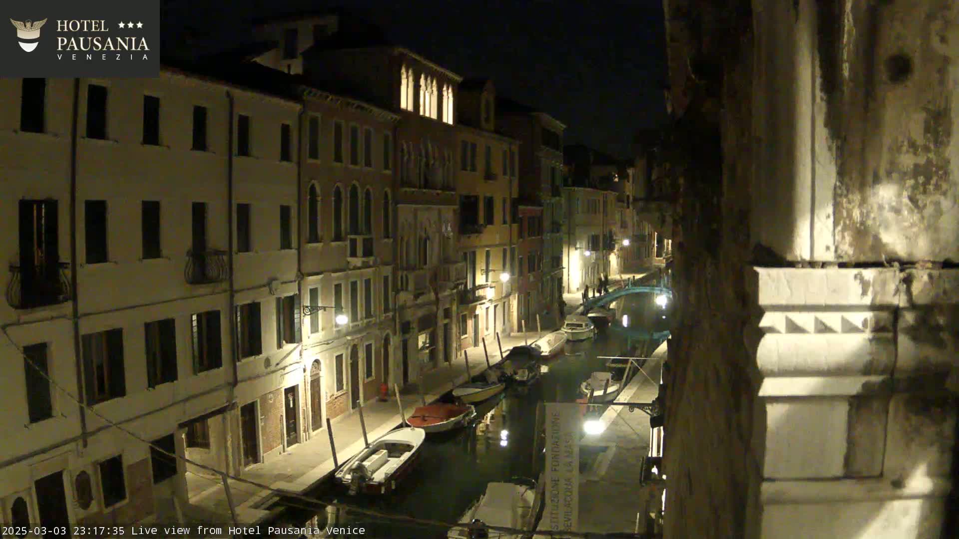 A nighttime view of a narrow canal in Venice, lined with illuminated buildings and several moored boats.