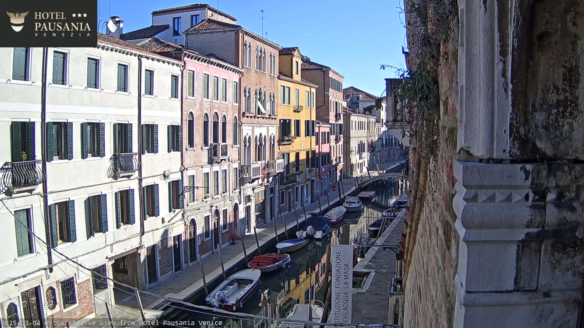 A sunny view down a narrow Venetian canal, lined with colorful buildings and several moored boats.