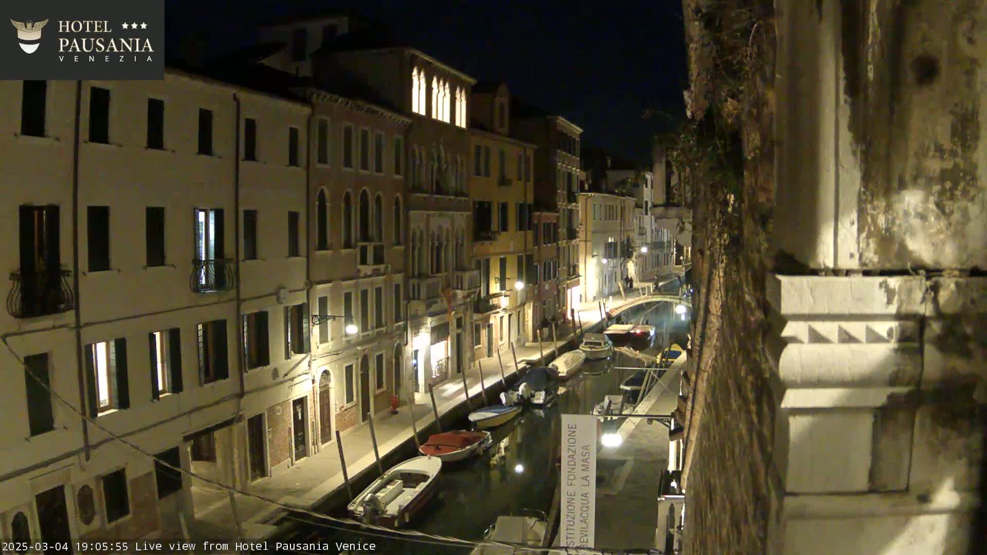 A nighttime view of a canal in Venice, Italy, lined with illuminated buildings and several moored boats.