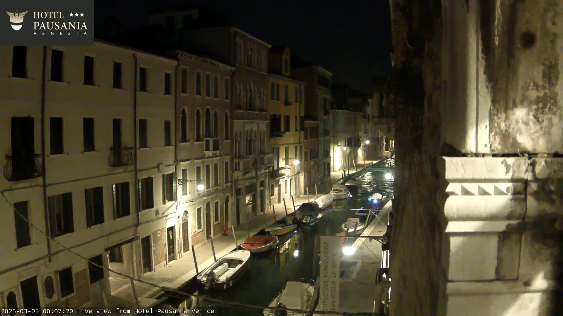 A nighttime view of a canal in Venice, showing several boats moored along the waterway, with illuminated buildings lining the canal banks.