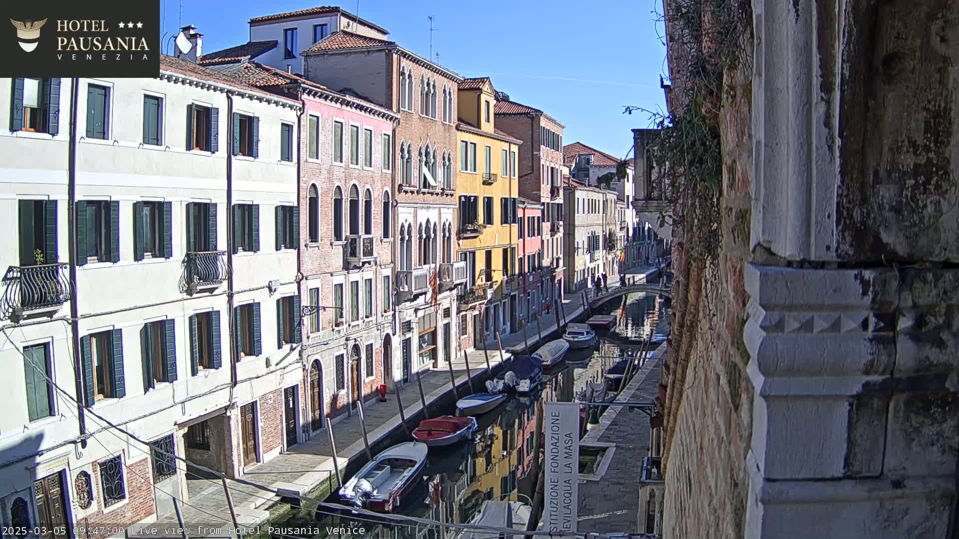 A sunny day in Venice shows several boats moored along a narrow canal, with colorful buildings lining the waterway.