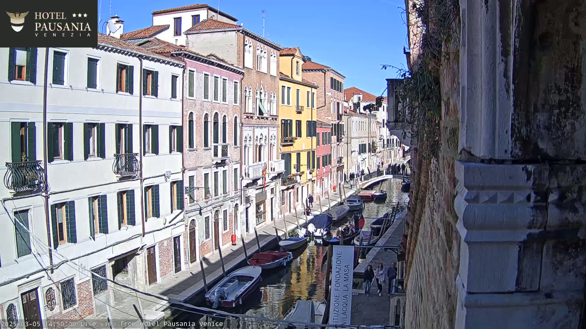 A sunny day in Venice shows a narrow canal lined with colorful buildings and several boats, some of which are moored along the waterway.