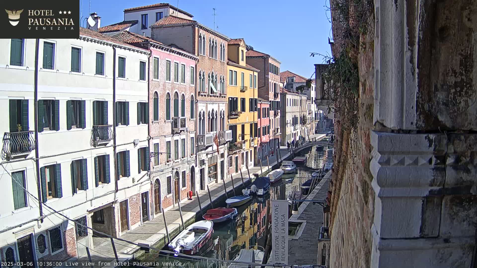 A sunny day in Venice shows several colorful buildings lining a canal with several boats moored along its edge.