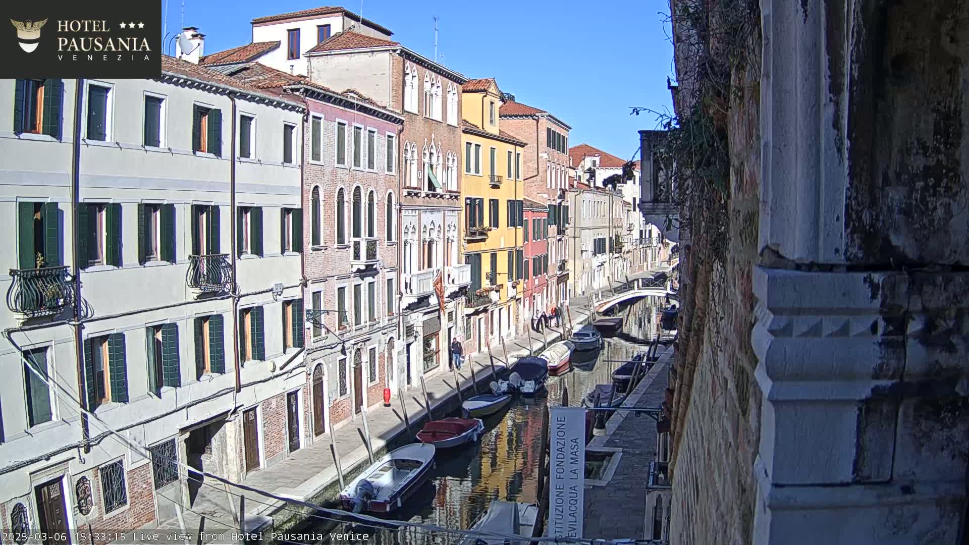 A sunny view of a narrow Venetian canal lined with colorful buildings and several boats moored along its banks.