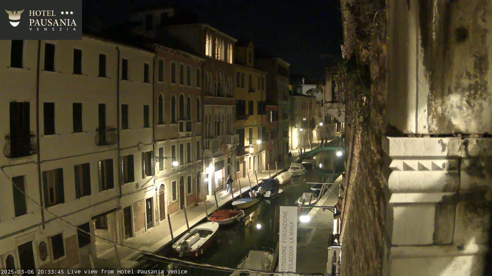 A nighttime view of a narrow Venetian canal lined with illuminated buildings and several moored boats.