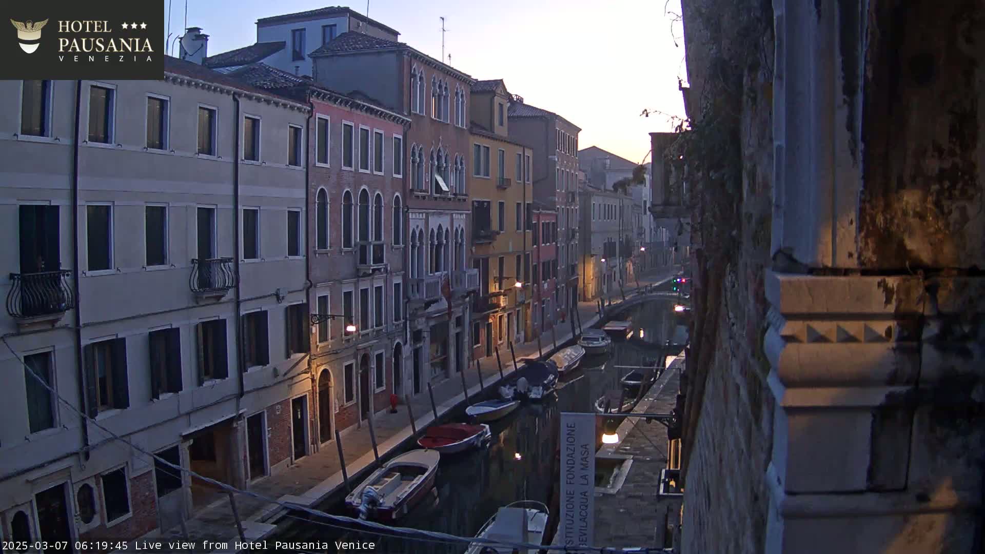 A twilight view of a narrow Venetian canal lined with multi-story buildings, several boats moored along the waterway.