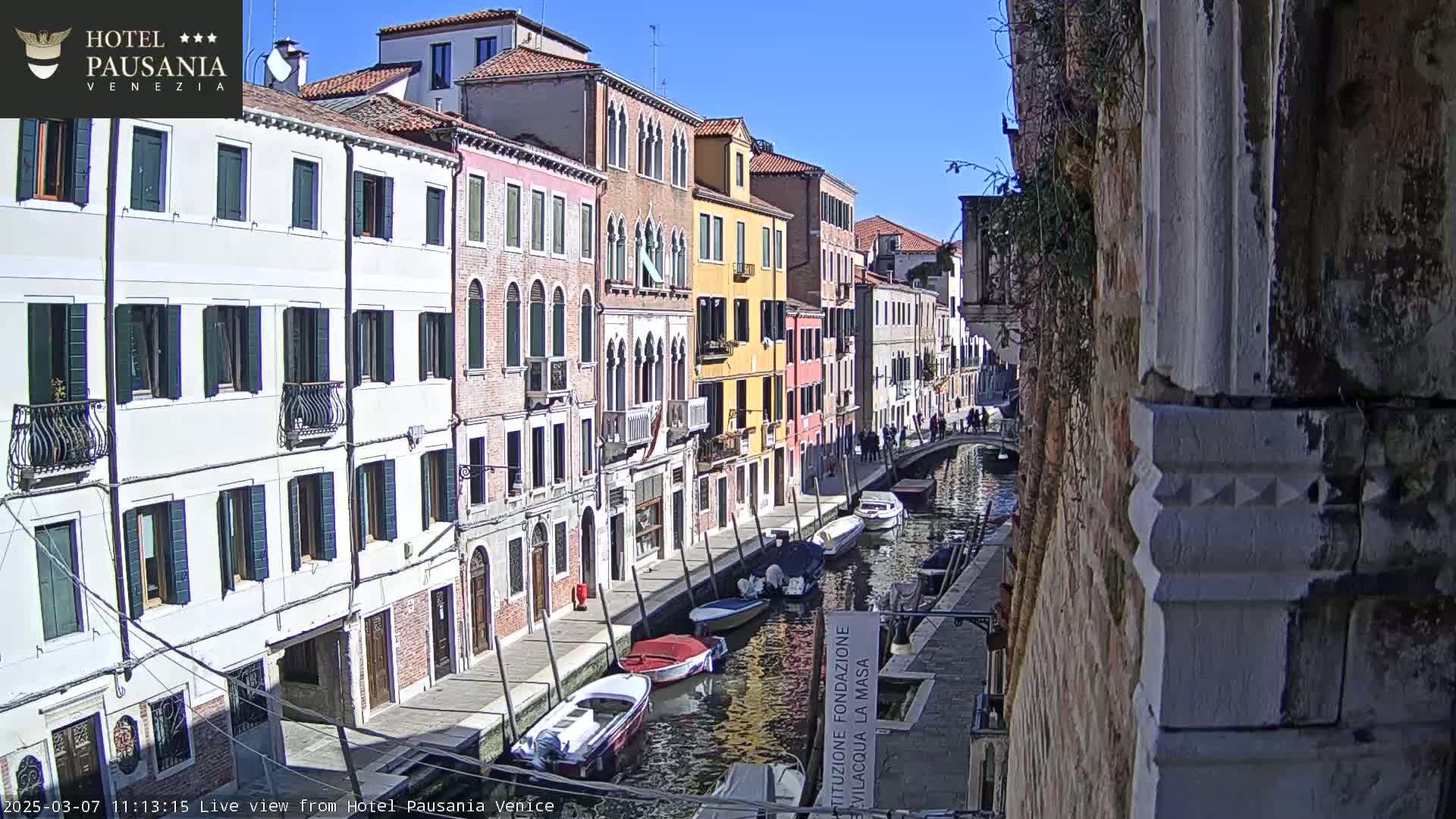 A sunny day in Venice shows colorful buildings lining a canal with several boats moored along its edge.