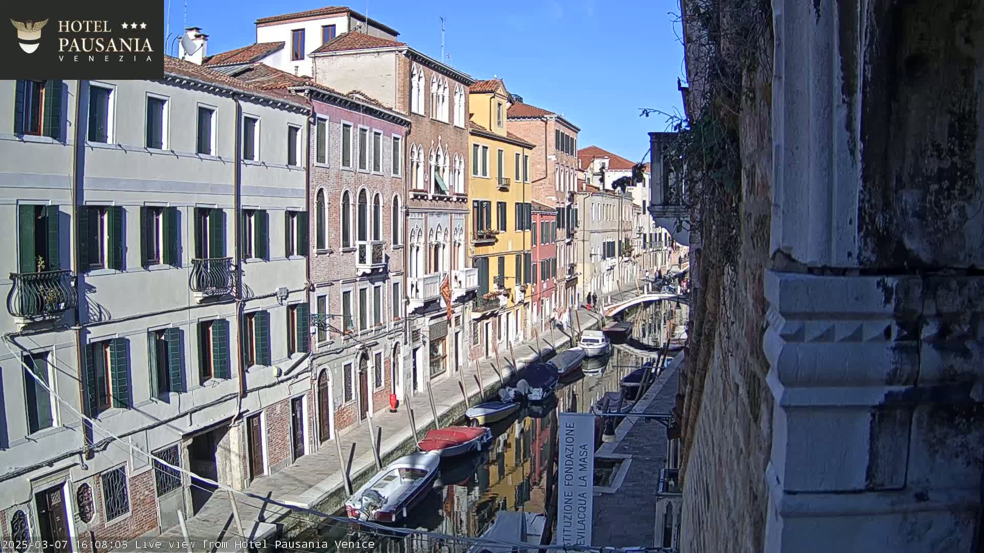 A sunny day in Venice shows a narrow canal lined with colorful buildings, several boats moored along its banks, and a stone wall partially obscuring the right side of the view.