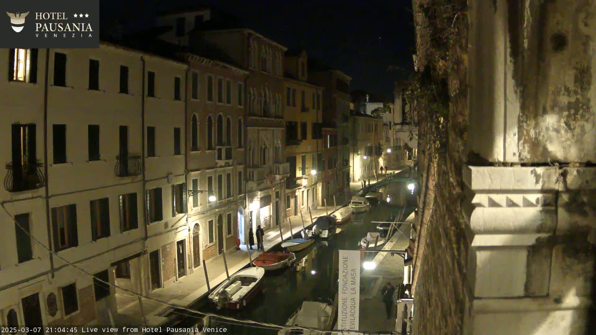 A nighttime view of a narrow canal in Venice, lined with illuminated buildings and several moored boats.