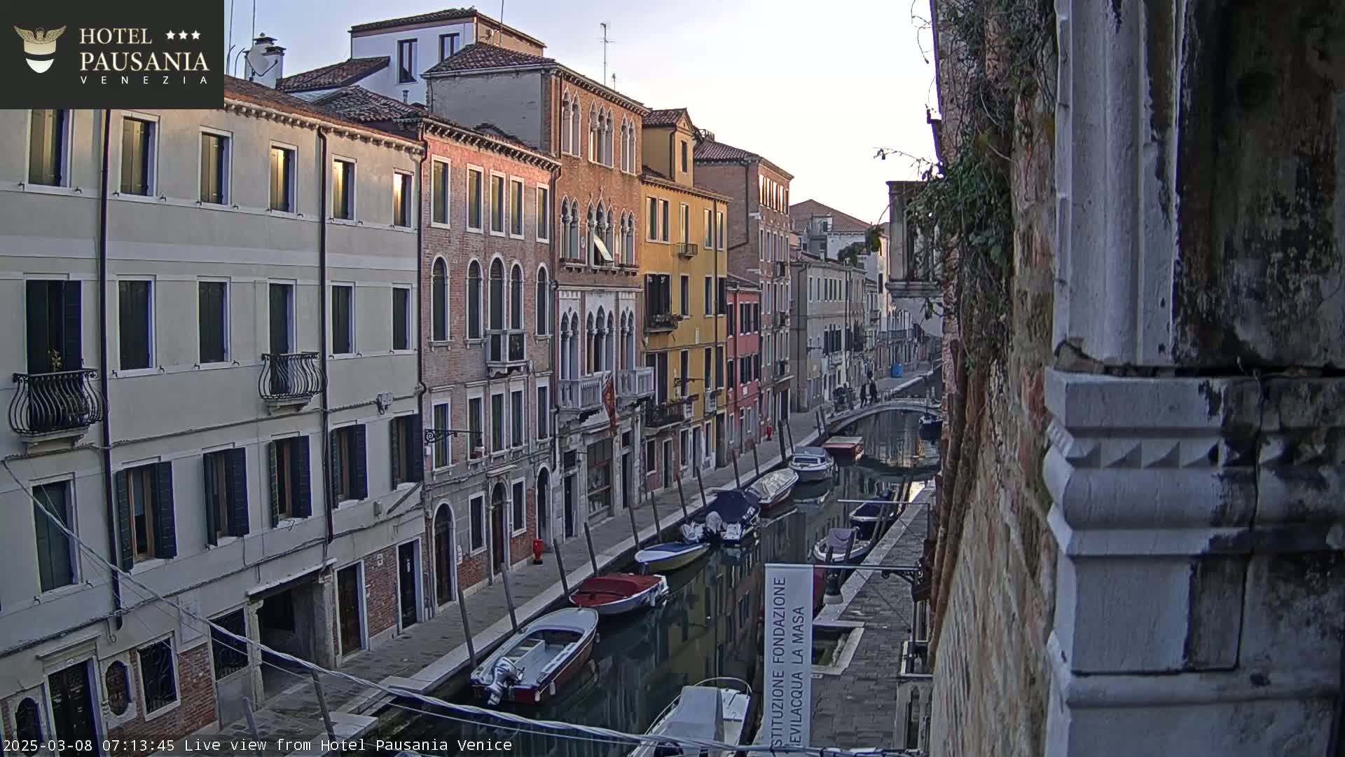 Several colorful buildings line a narrow canal in Venice, with several small boats moored along the waterway, under a clear, dawn sky.