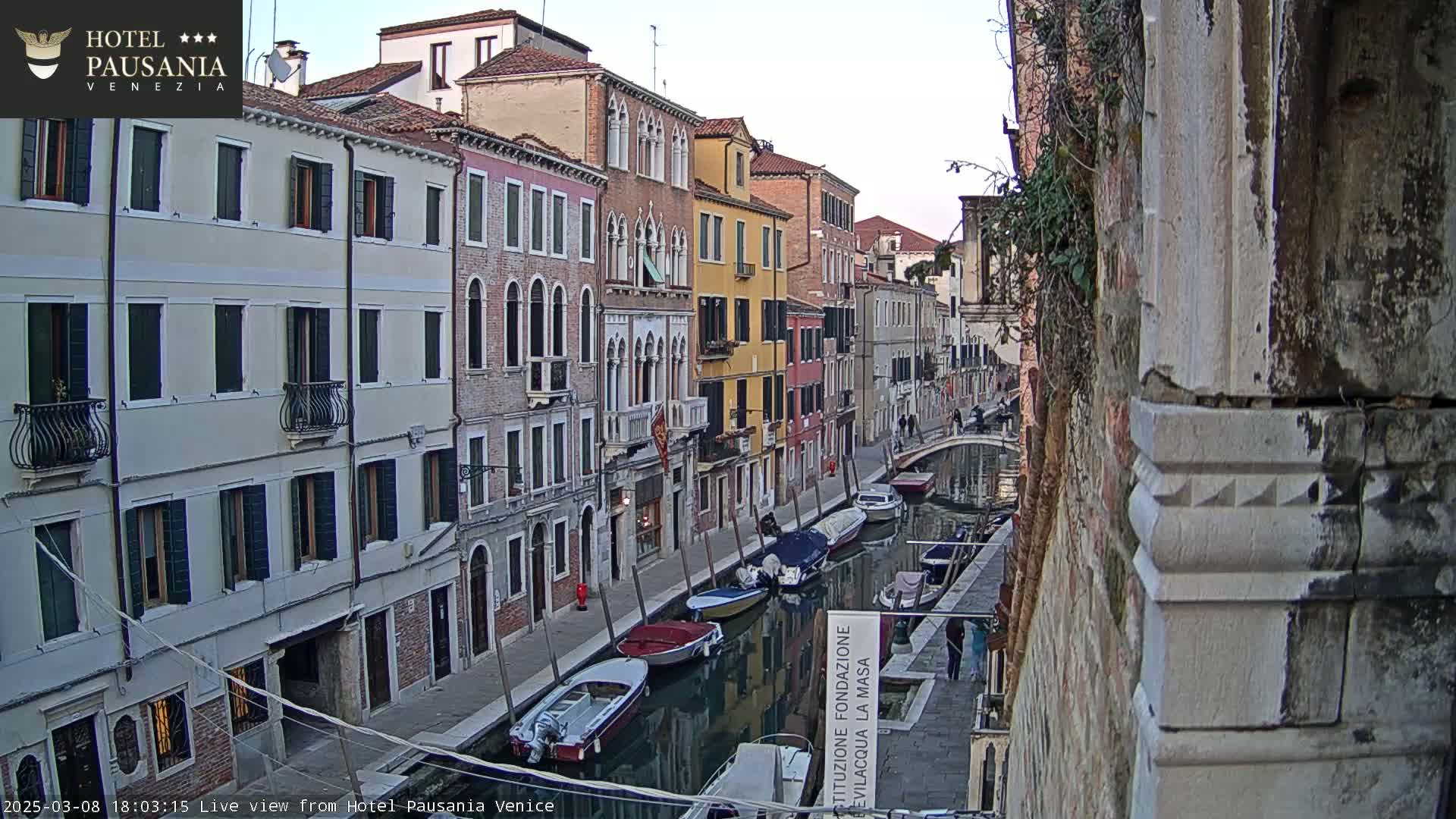 A narrow Venetian canal at dusk is lined with closely spaced multi-story buildings, and several small boats are moored along its banks.