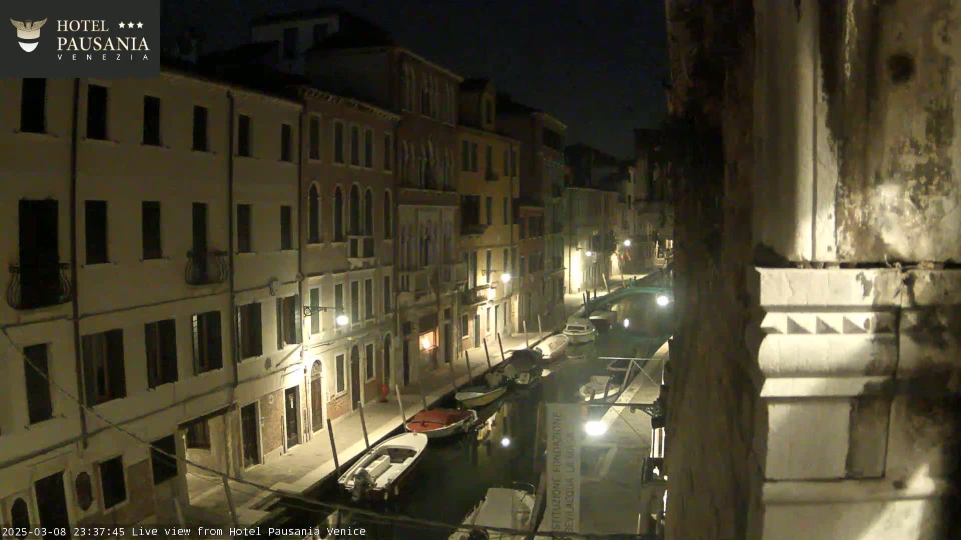 A nighttime view of a narrow canal in Venice, lined with buildings and moored boats, under a dark sky.
