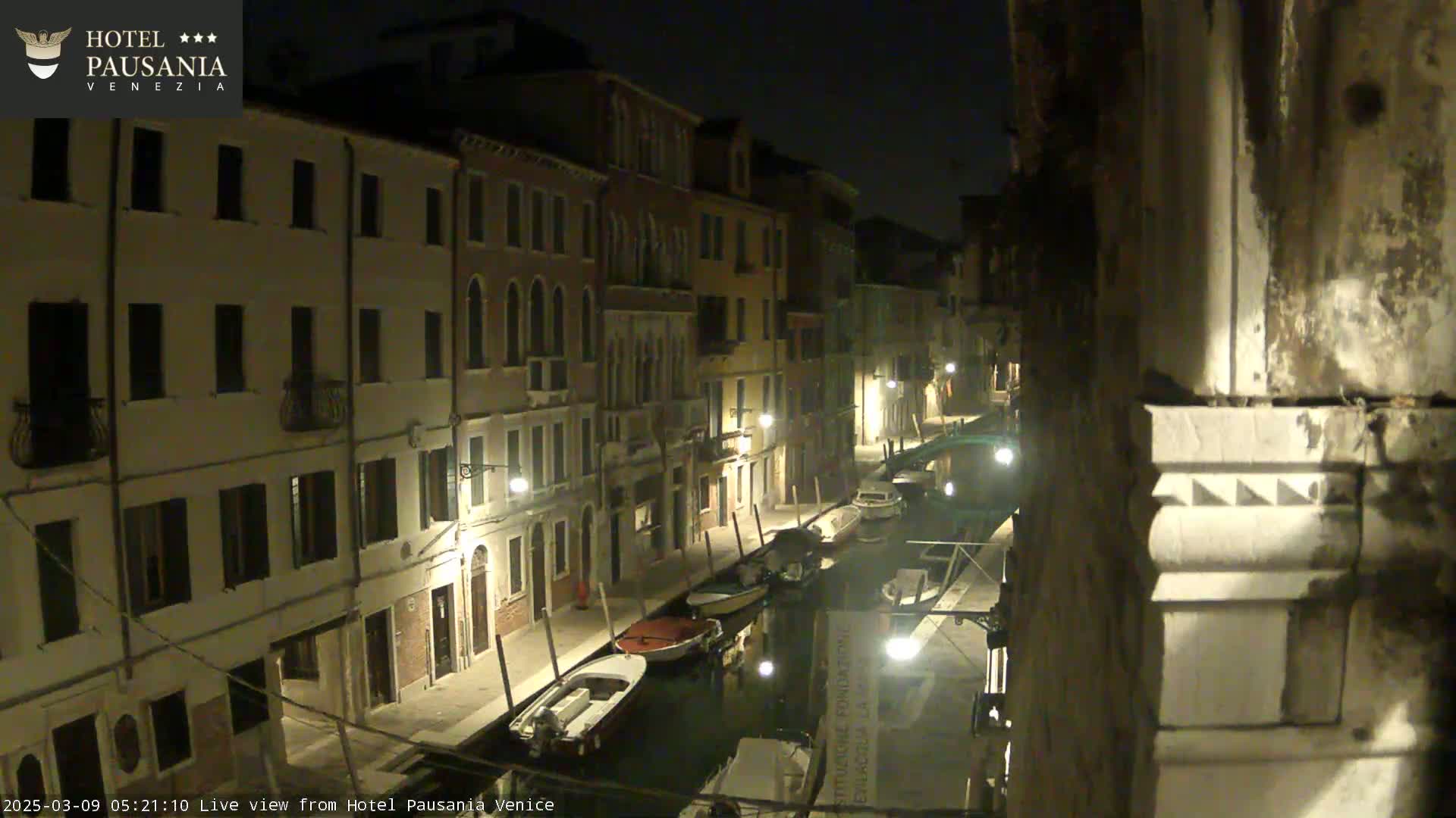 A nighttime view of a narrow canal in Venice, with several boats moored alongside buildings illuminated by streetlights.