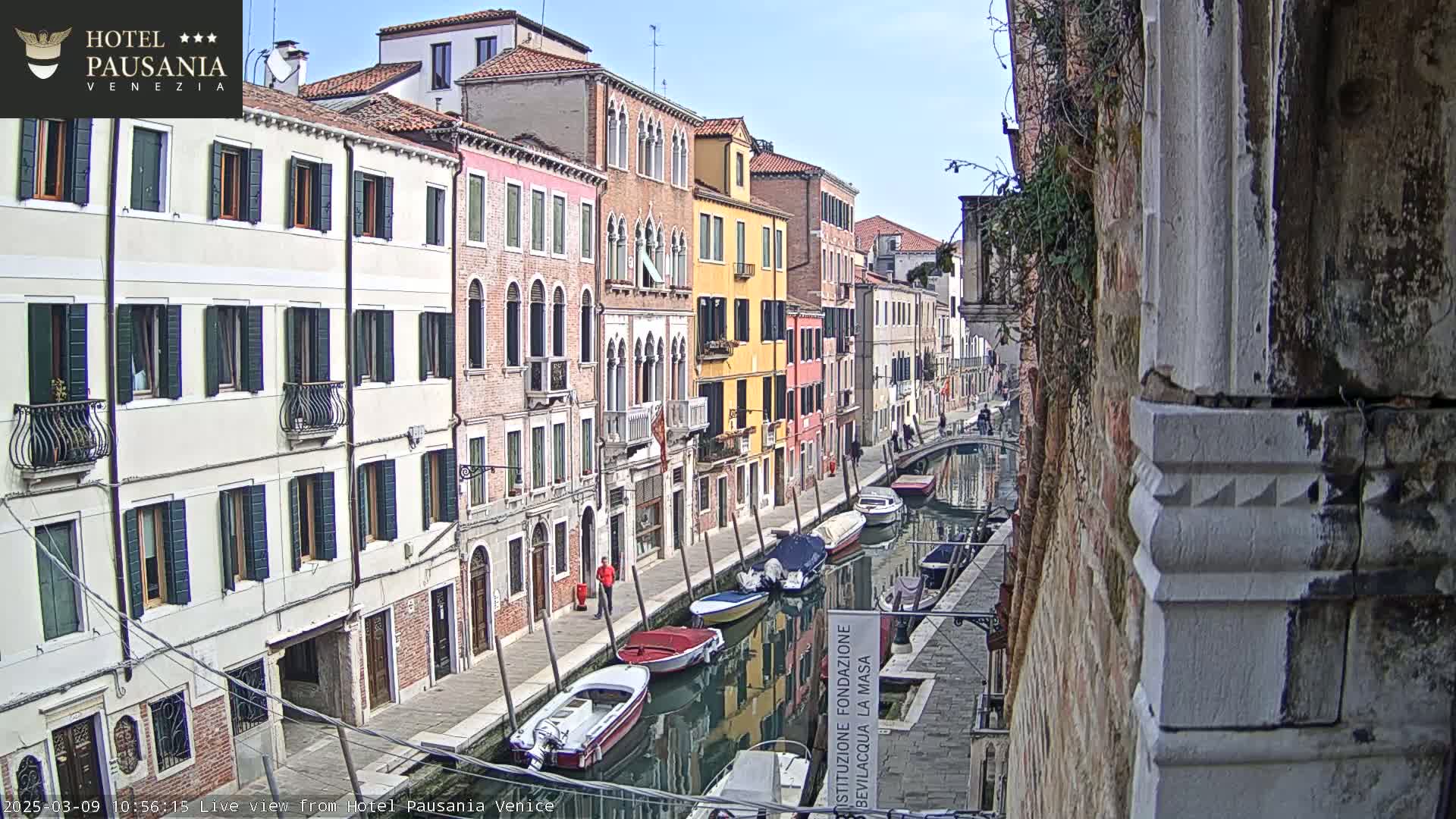 A narrow Venetian canal, lined with colorful buildings, under a mostly clear sky, shows several boats moored along its sides.
