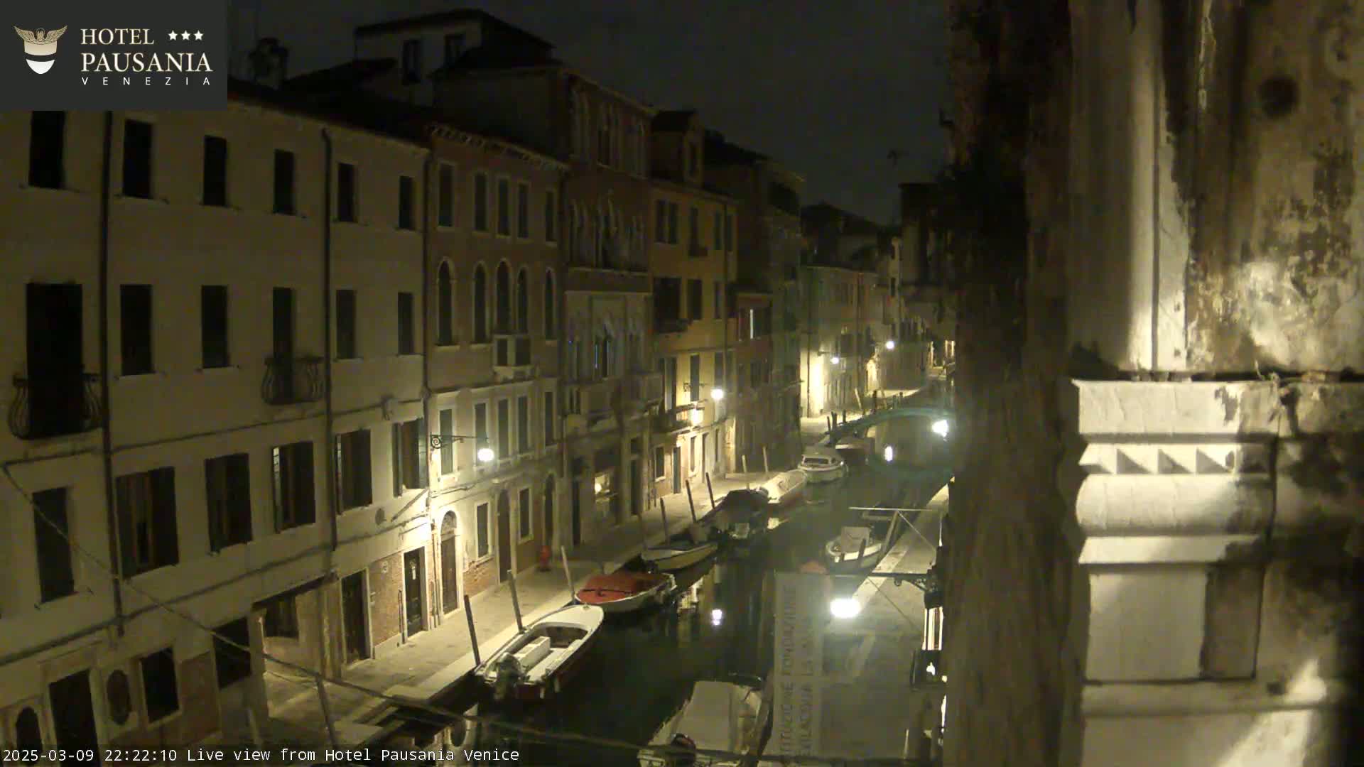 A nighttime view of a narrow canal in Venice, lined with buildings and moored boats, under a clear, dark sky.