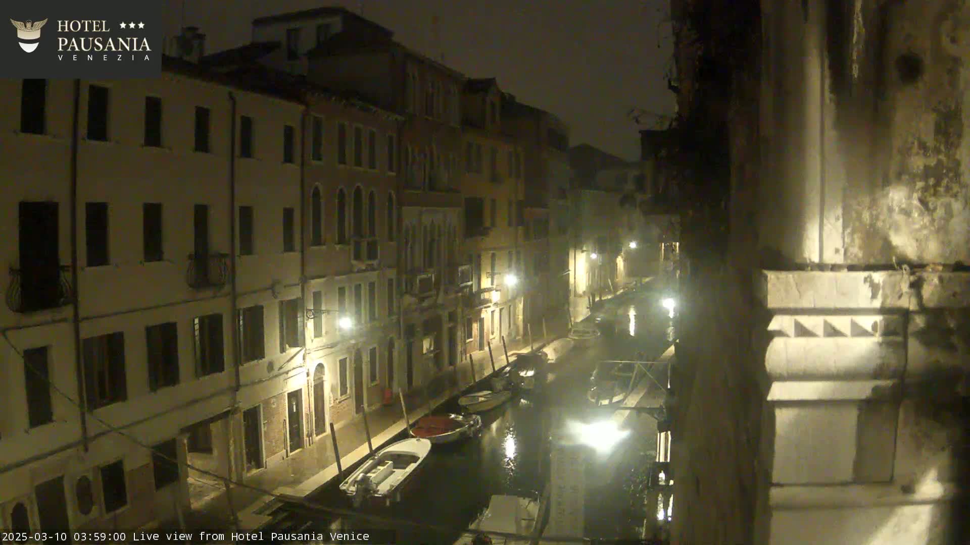 A nighttime view of a canal in Venice, showing several buildings lining the waterway and a few boats moored along the canal's edge, under a dark and possibly misty sky.