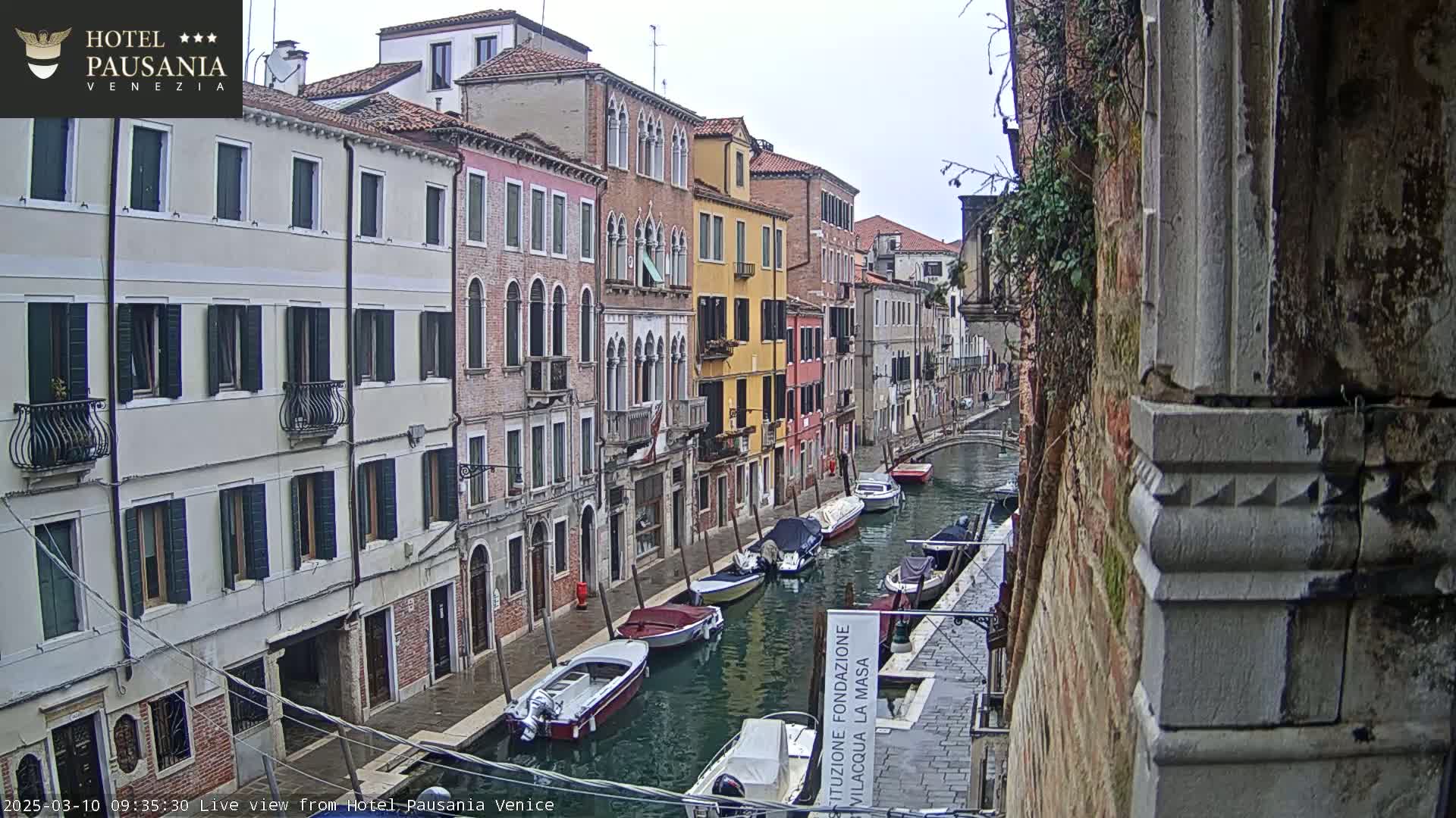 Several boats are moored along a narrow canal in Venice, flanked by tall, colorful buildings under an overcast sky.