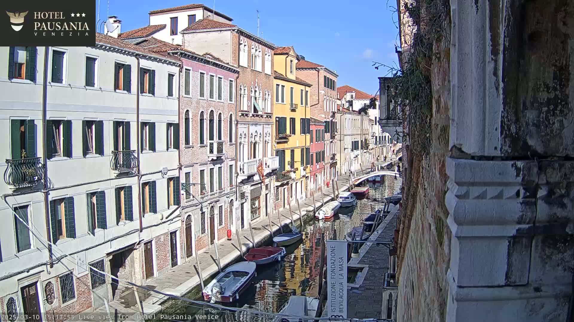 A sunny day in Venice shows several boats moored along a narrow canal, flanked by colorful buildings.