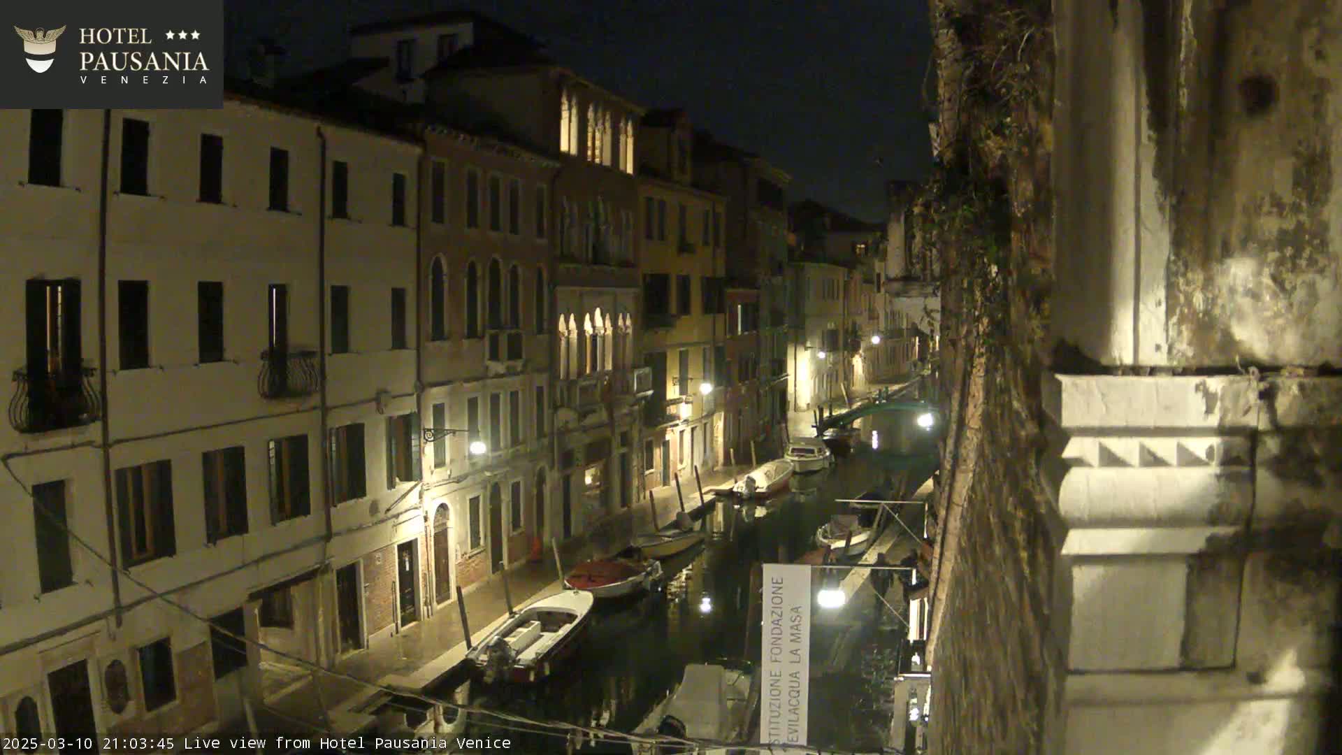 A nighttime view of a narrow canal in Venice, lined with illuminated buildings and several moored boats.