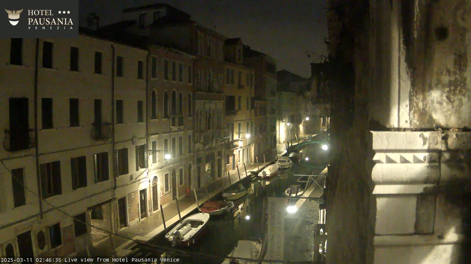 A nighttime view of a narrow canal in Venice, Italy, lined with buildings and several moored boats.