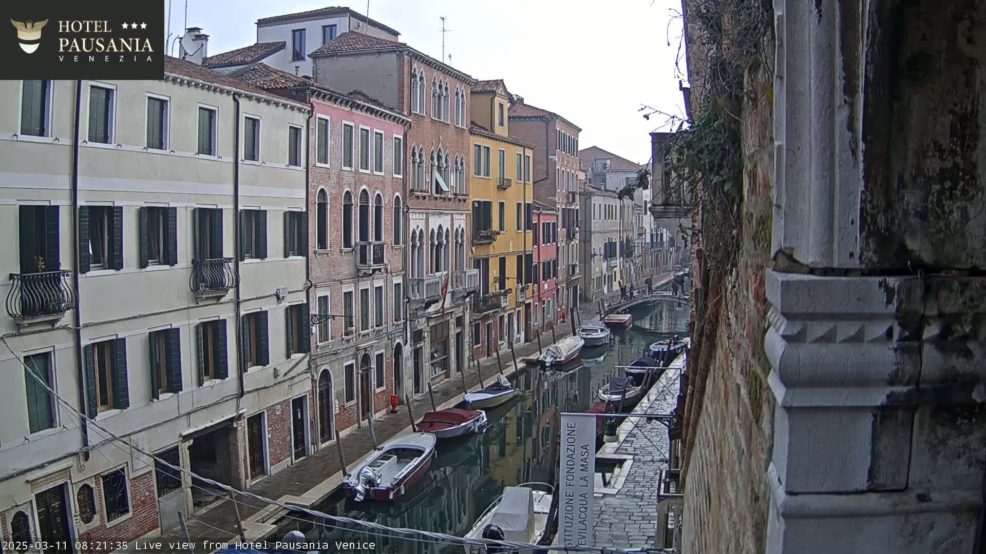 Several moored boats are in a narrow canal lined with closely-spaced, multi-storied buildings on a cloudy day.