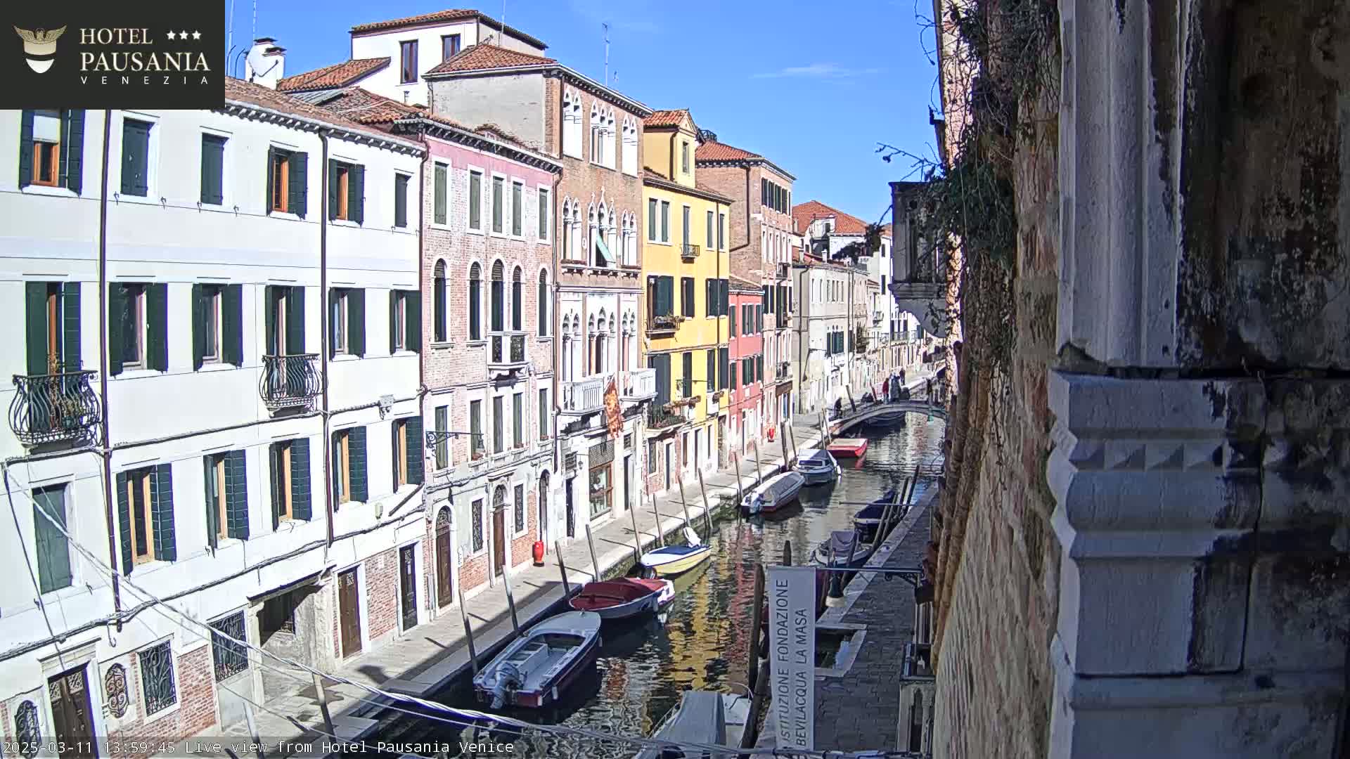 A sunny day in Venice shows colorful buildings lining a canal with several boats moored along its edge.