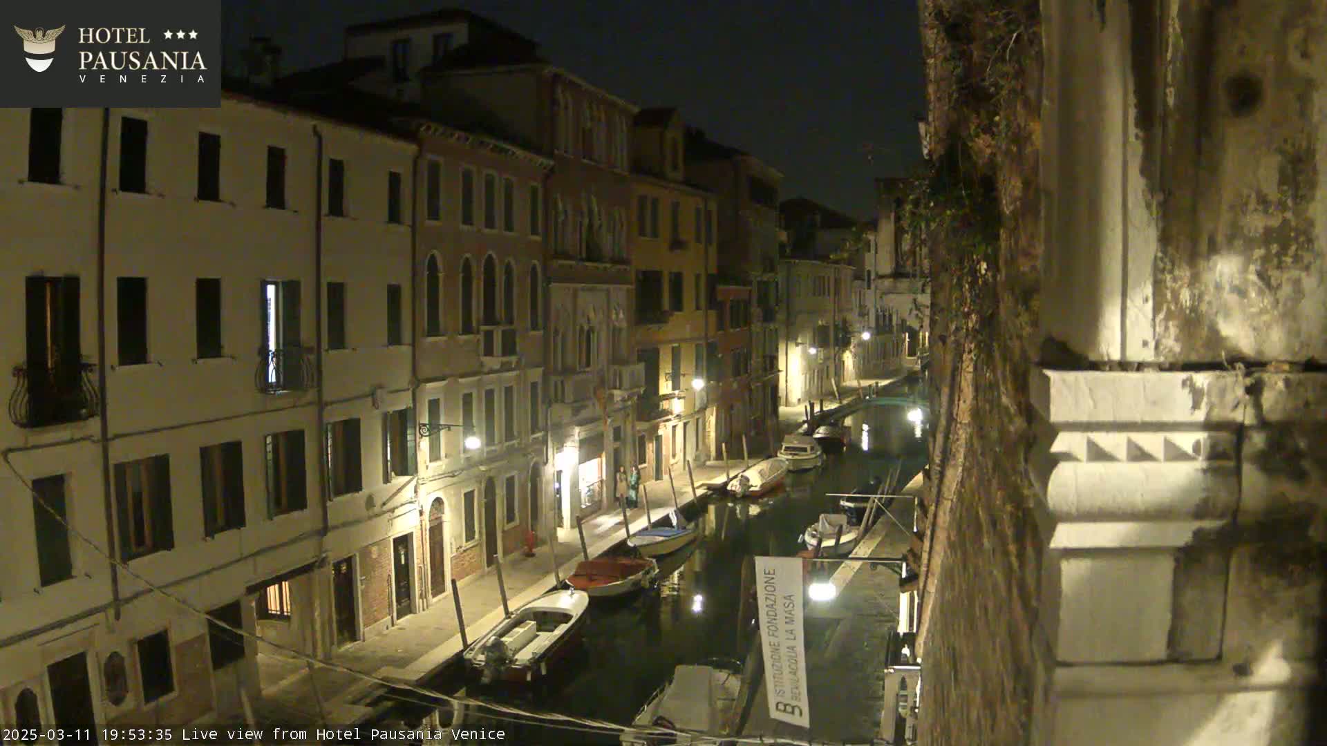 At night, several boats are moored along a canal lined with illuminated buildings.