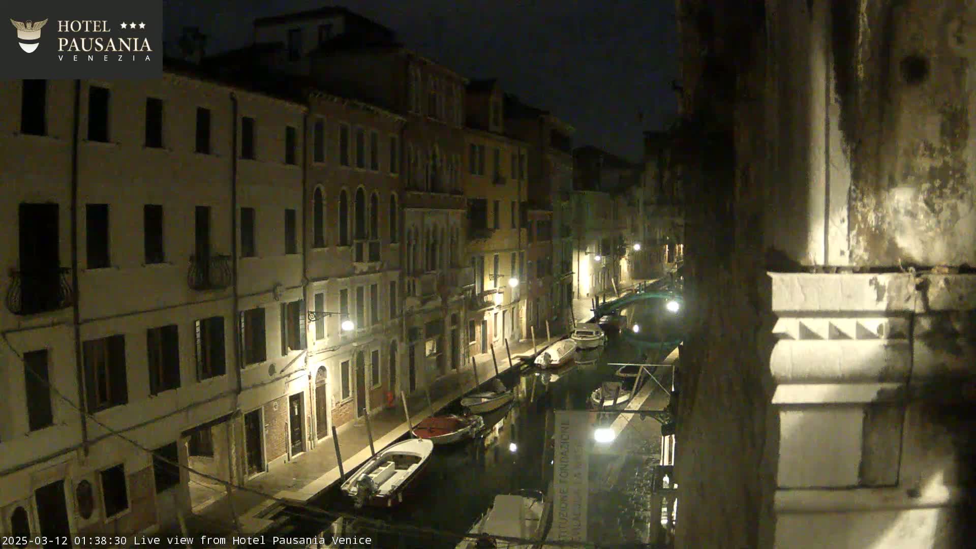 A nighttime view of a narrow canal in Venice, showing several boats moored along the waterway and buildings lining the sides, under a dark sky.