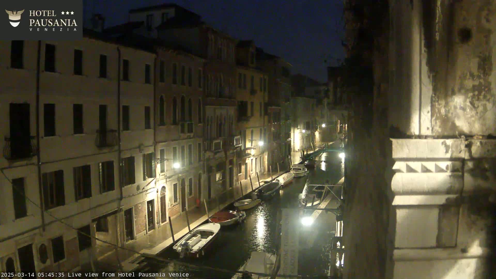A nighttime view of a narrow canal in Venice, lined with buildings and several moored boats, under a dark sky.