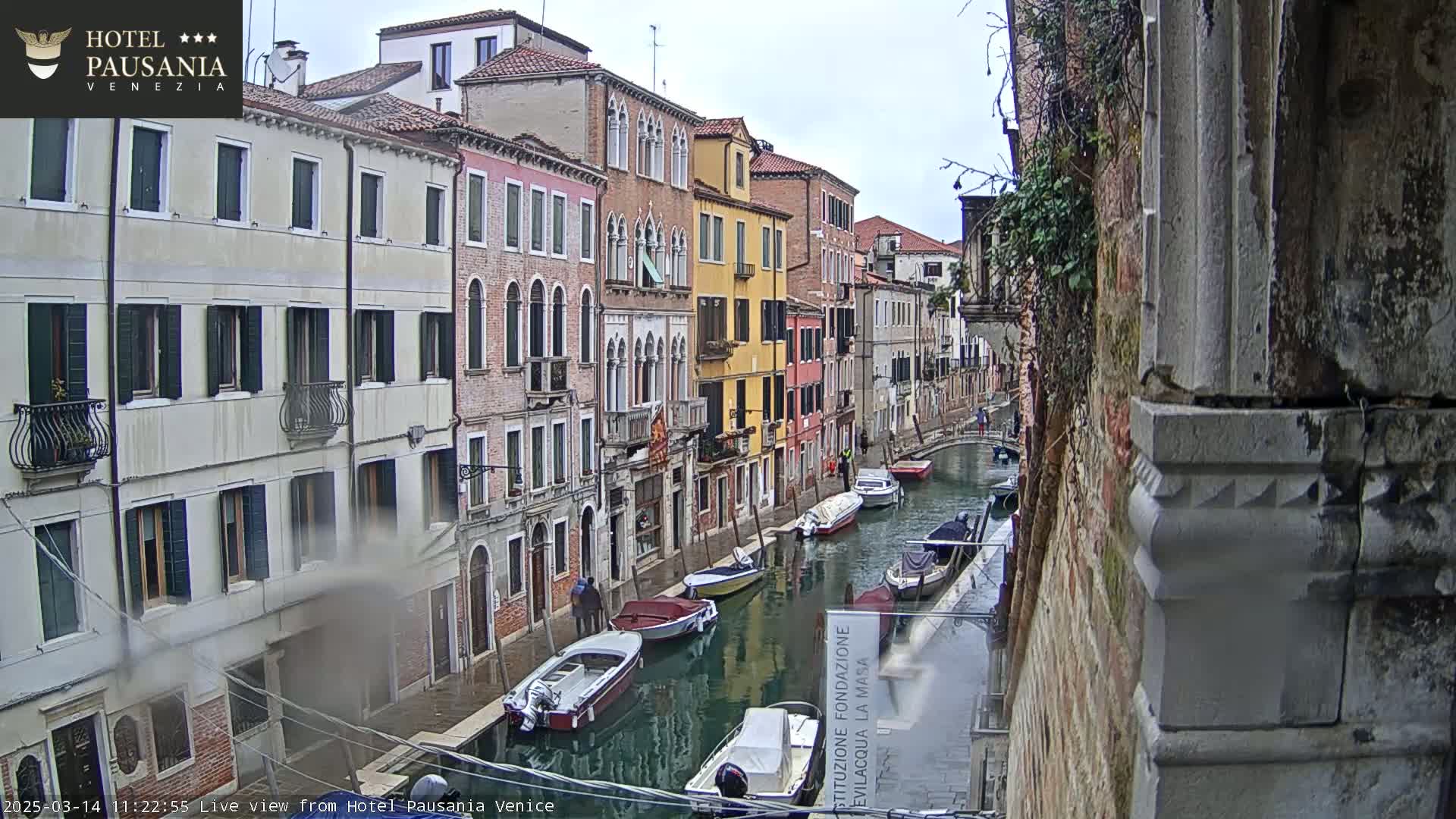 A narrow Venetian canal is lined with colorful buildings under an overcast sky, several small boats are moored along its banks.