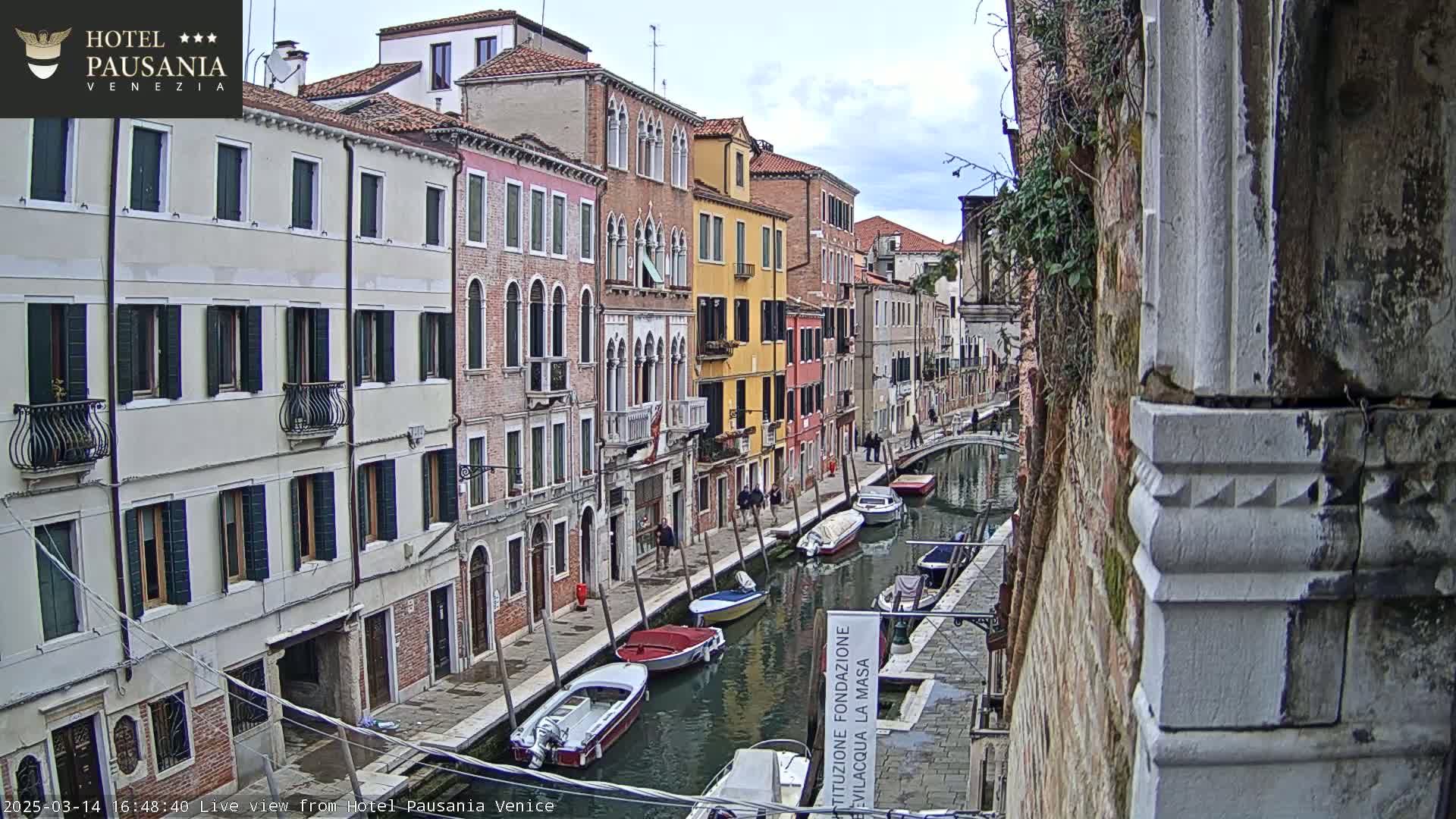 Overcast skies loom over a narrow Venetian canal, lined with colorful historic buildings and several moored boats.