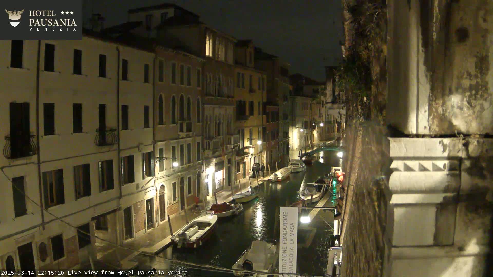 A nighttime view of a narrow canal in Venice, lined with illuminated buildings and several moored boats.