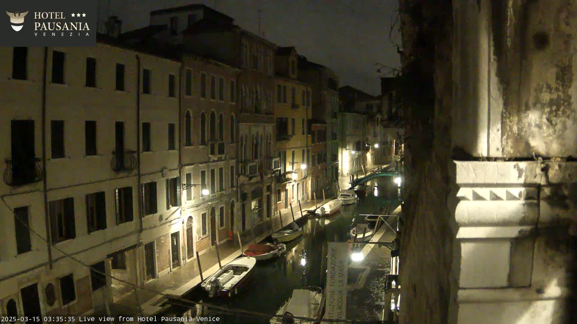 A nighttime view of a canal in Venice, showing several moored boats and buildings lining the waterway under dimly lit streetlights.