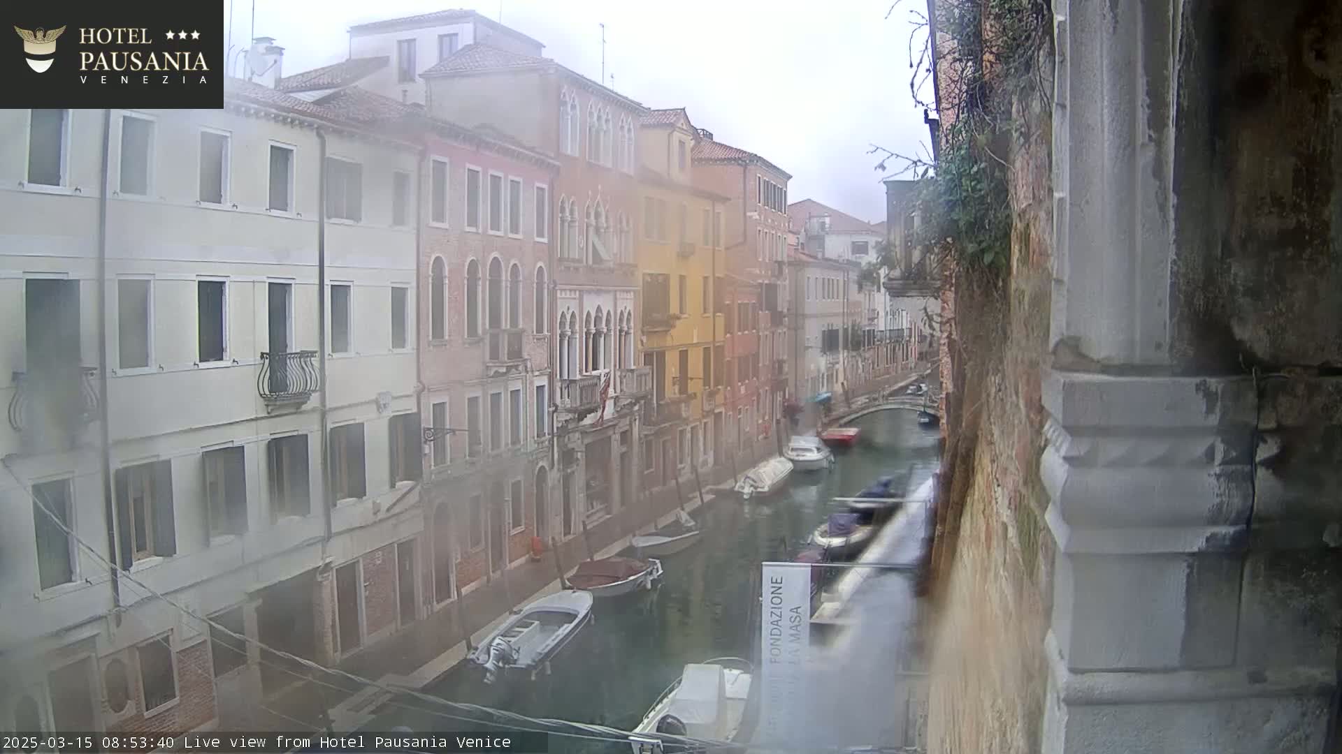 A hazy view of a narrow Venetian canal, with several boats moored along its sides, on an overcast day.