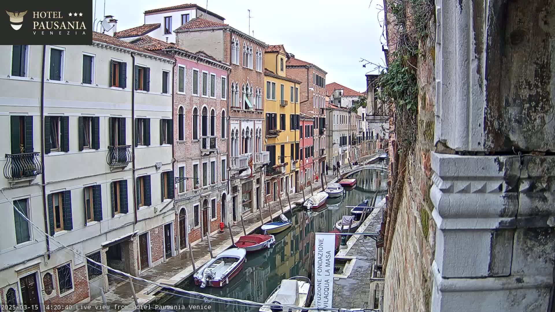 A narrow canal in Venice, Italy, is lined with colorful buildings under an overcast sky, and several boats are moored along its banks.