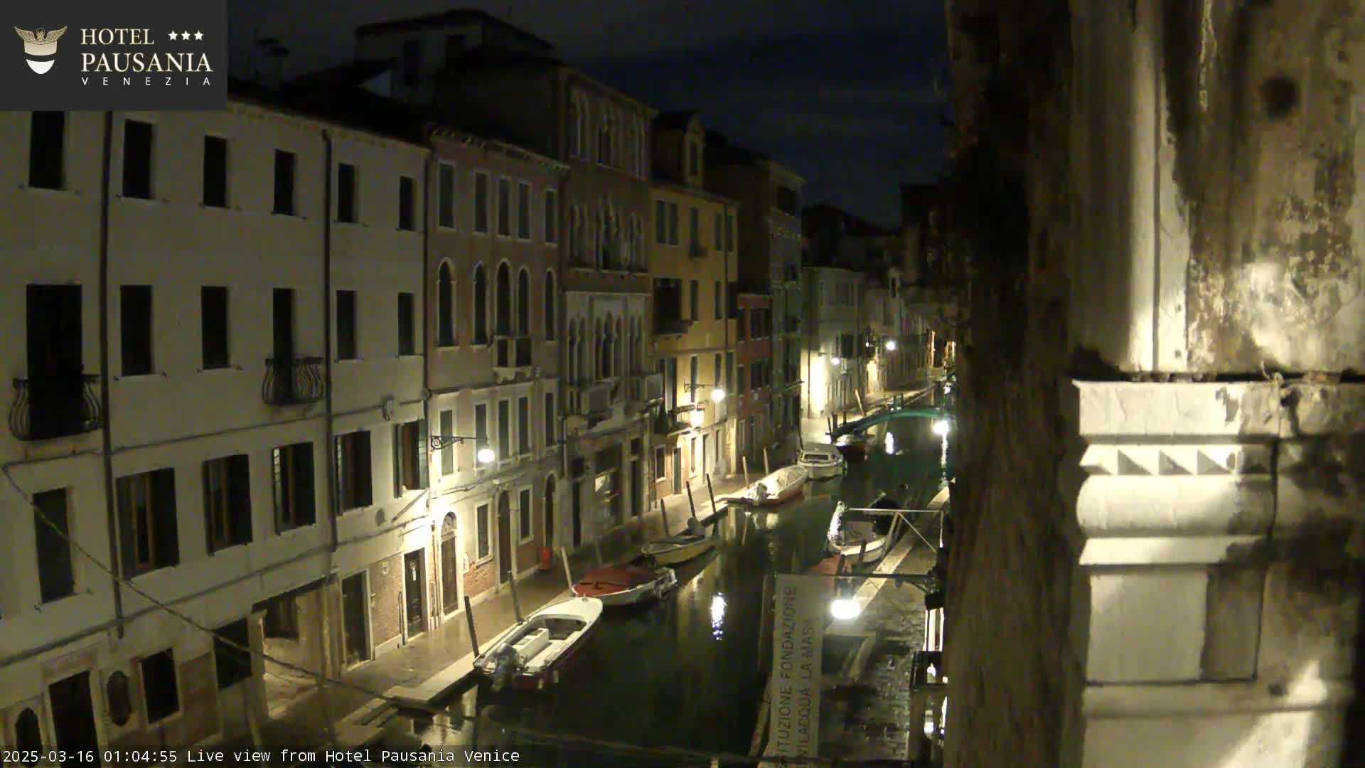 A nighttime view of a narrow canal in Venice, lined with buildings and several moored boats under a dark sky.