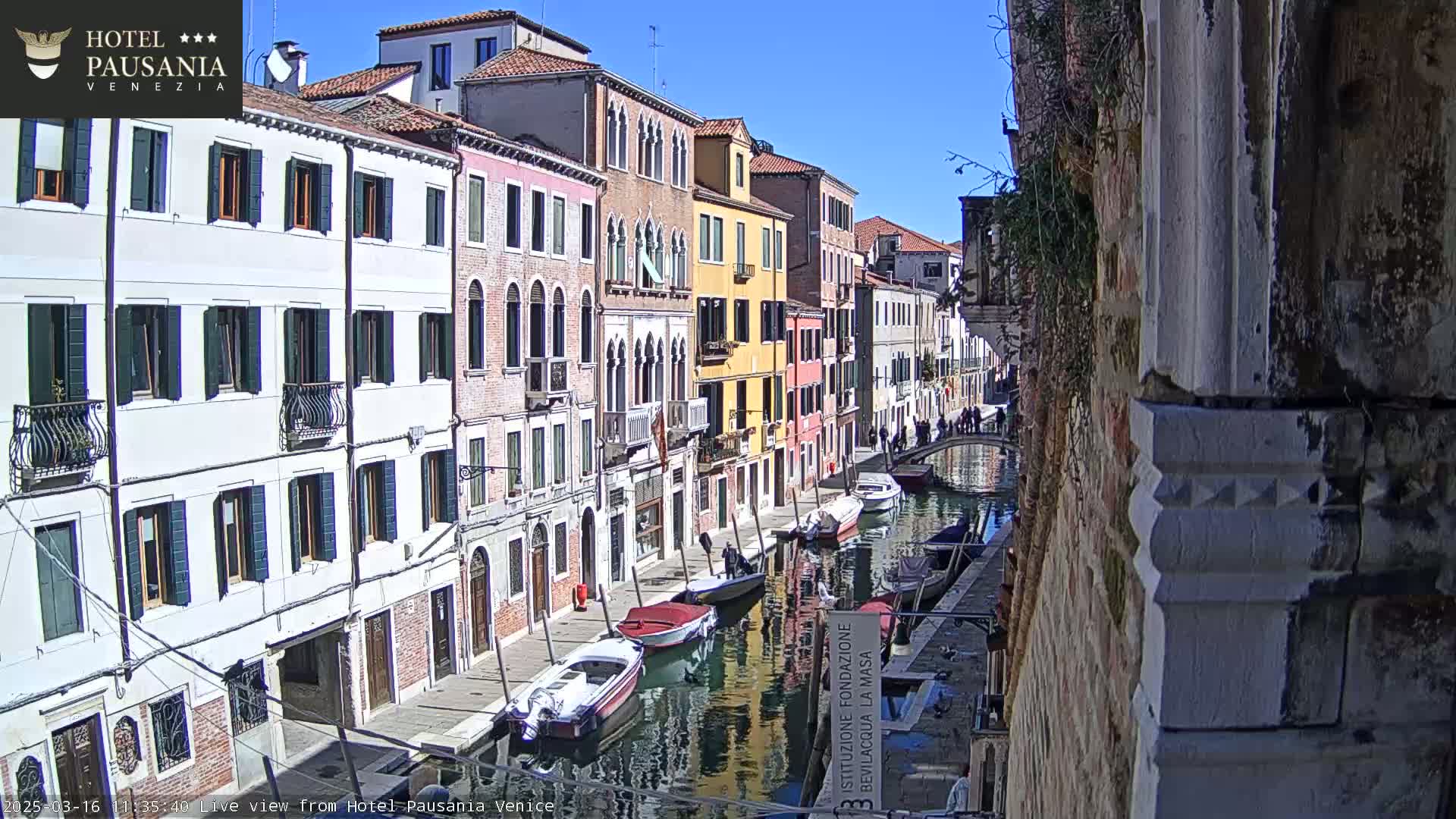 A sunny day in Venice shows colorful buildings lining a canal with several boats moored along its edge.