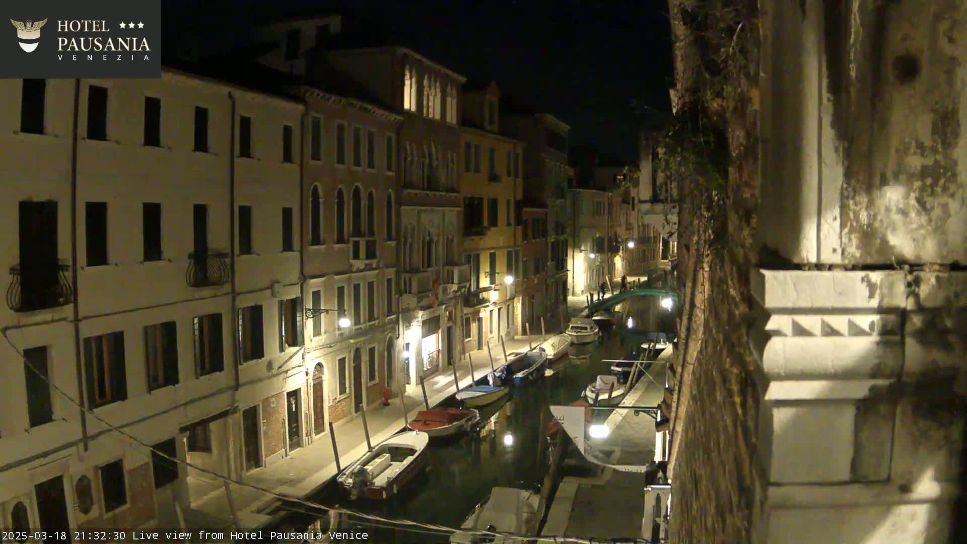 A nighttime view of a narrow canal in Venice, lined with buildings and several moored boats.