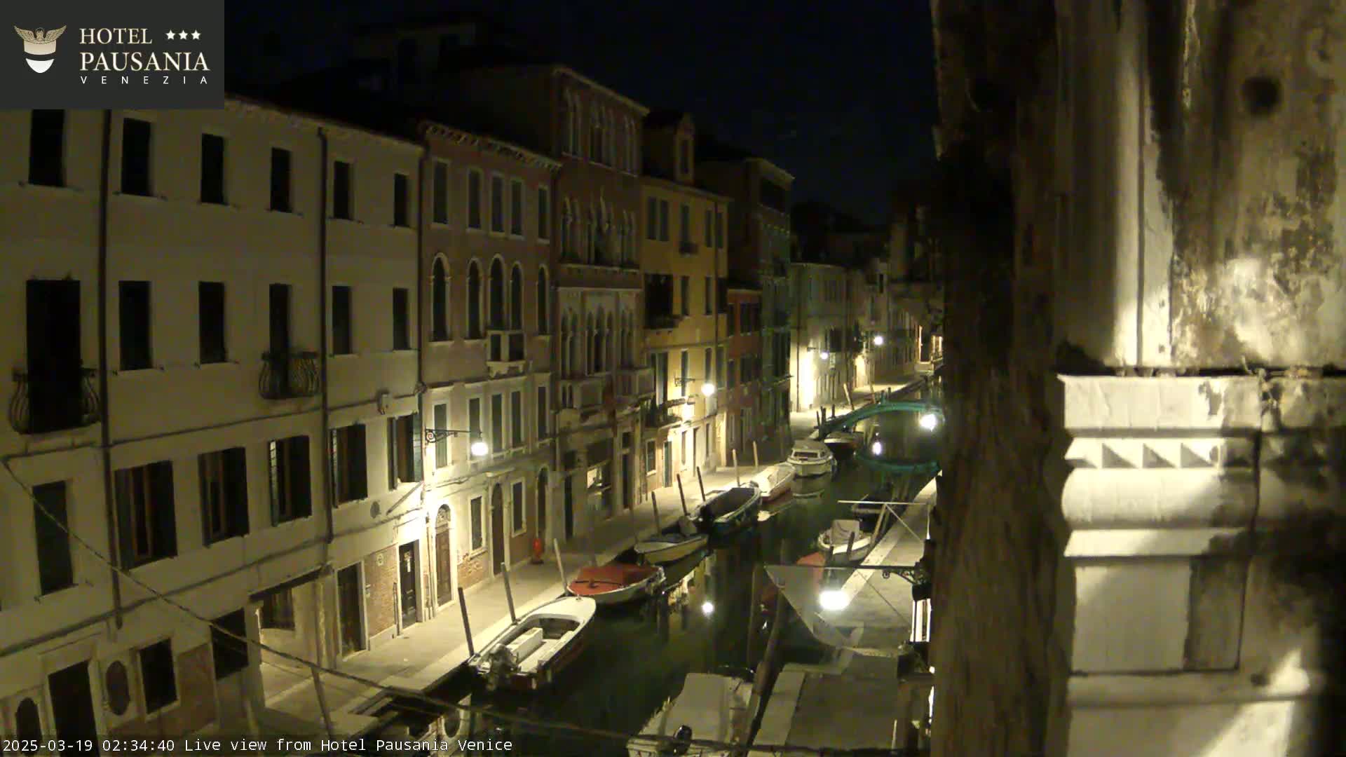 A nighttime view of a narrow canal in Venice, Italy, lined with illuminated buildings and several moored boats.
