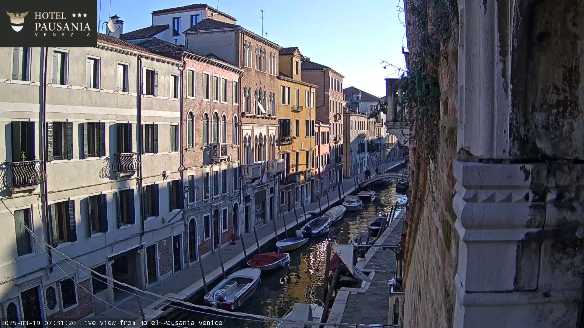 A sunny view down a narrow canal in Venice, lined with closely spaced buildings, showing several boats moored along the waterway.